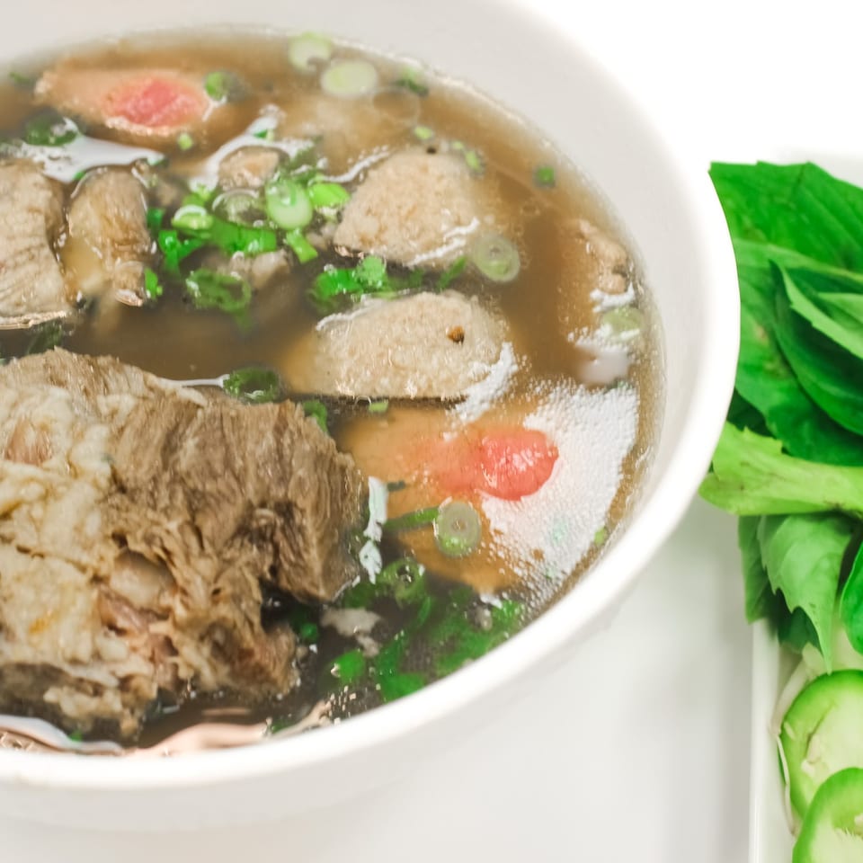 A bowl of pho with beef, meatballs, and green onions, served with a side of fresh herbs.
