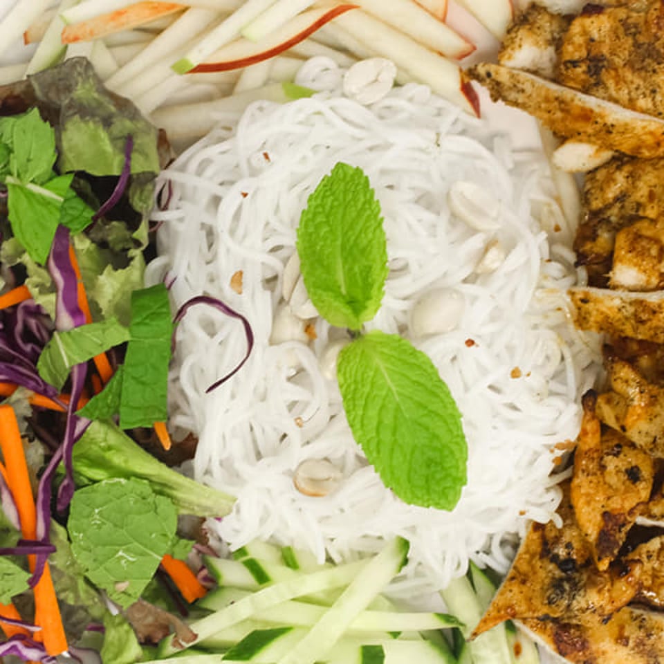 Grilled chicken, vermicelli noodles, fresh mint, lettuce, carrots, red cabbage, and cucumber on a white plate.